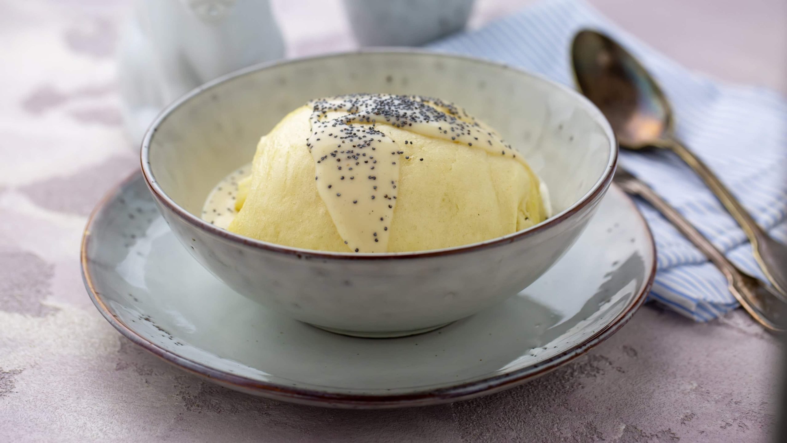 Eine Dampfnudel in einem weißen tiefen Teller auf einem Steintisch mit Löffeln.