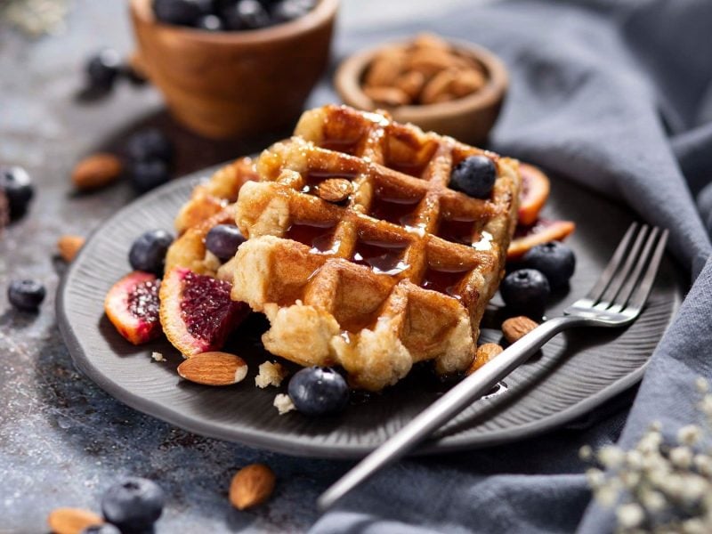 Draufsicht: Haferkleie-Waffeln mit Beeren und Mandeln liegen auf einem dunklen Teller auf einem dunklen Untergrund.