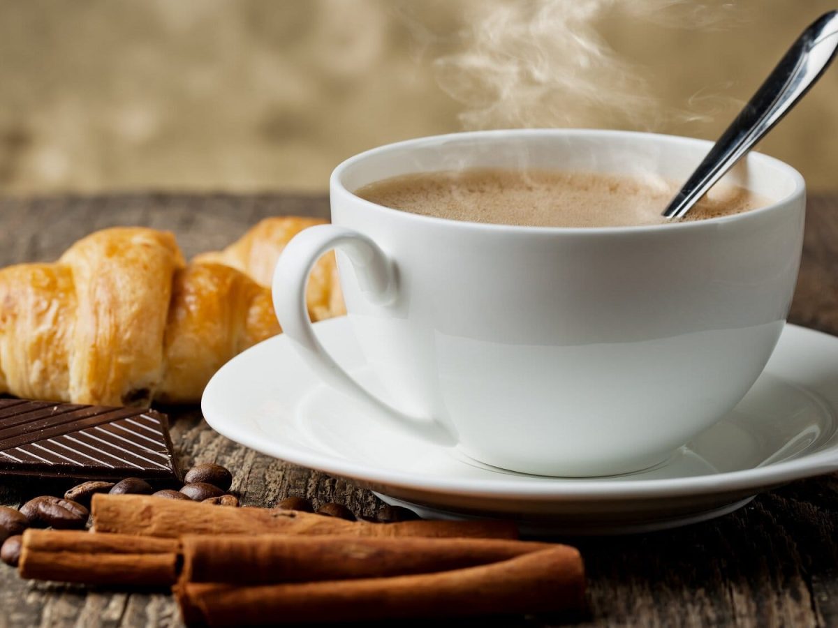 Frontalsicht: Ein Kaffee mit Vanille steht auf einem Holztisch. Daneben liegen Vanilleschoten und ein Croissant.