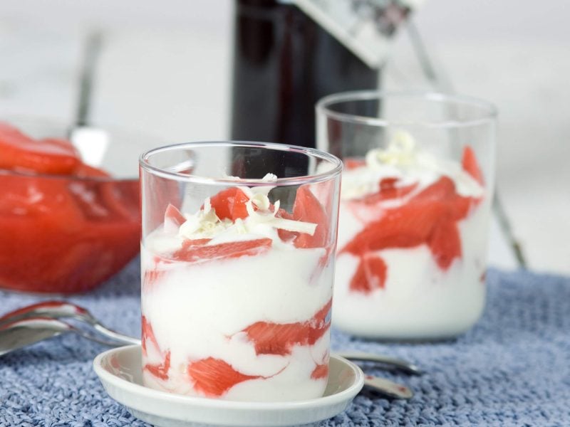 Frontalsicht: Zwei Gläser Rhabarber-Joghurt. Dahinter steht eine Schüssel aus Glas mit Rhabarber-Kompott.