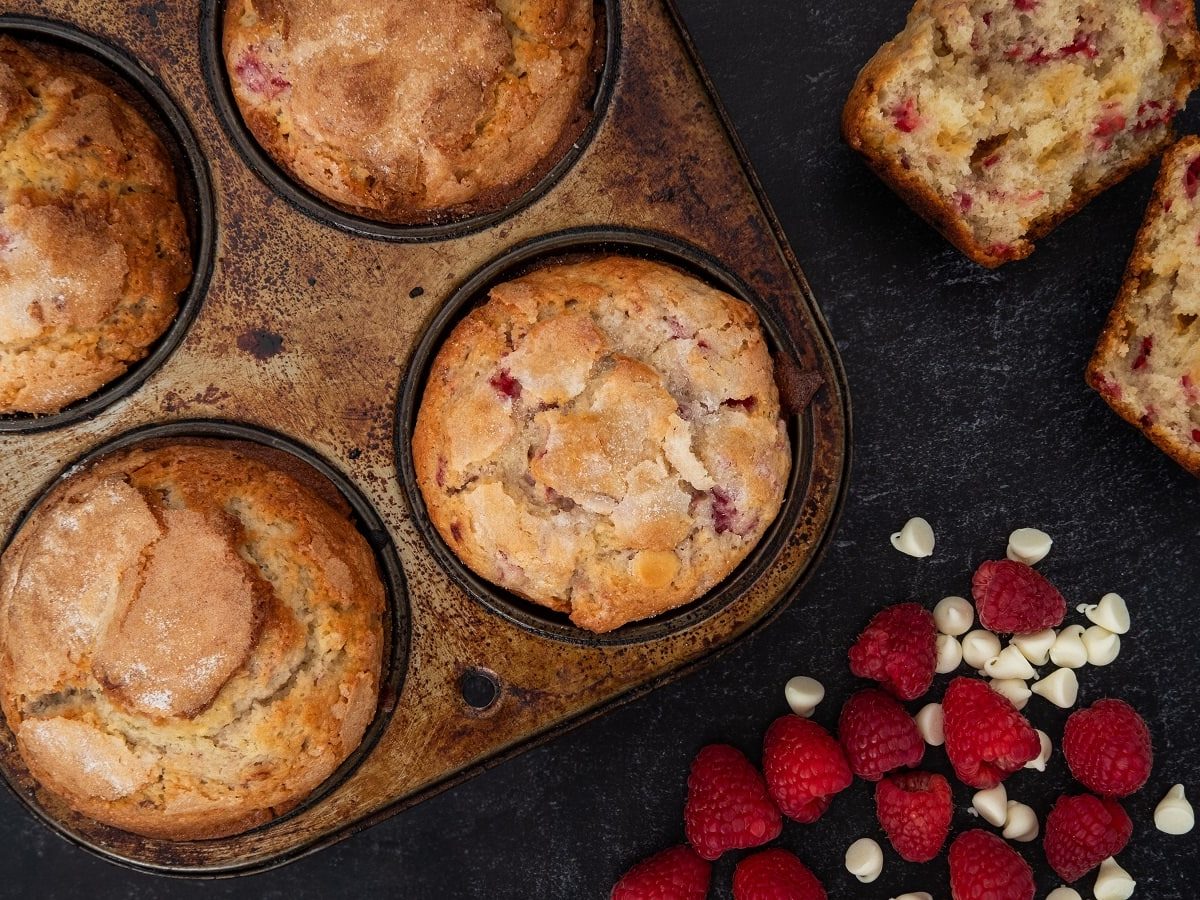 Ein Blech mit Himbeer-Muffins mit weißer Schokolade auf dunklem Hintergrund.