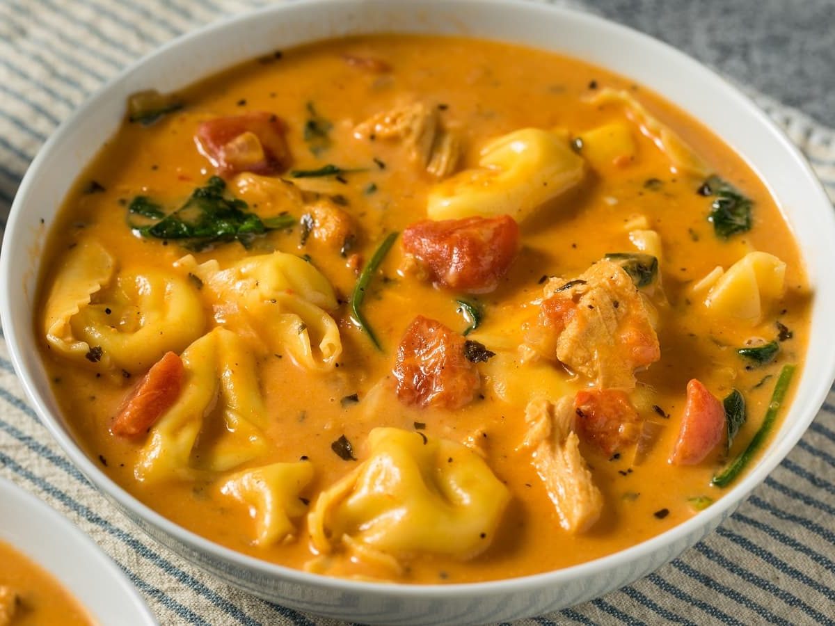 Eine weiße Schüssel Tortellini-Tomatencremesuppe auf einem hellen Tisch.