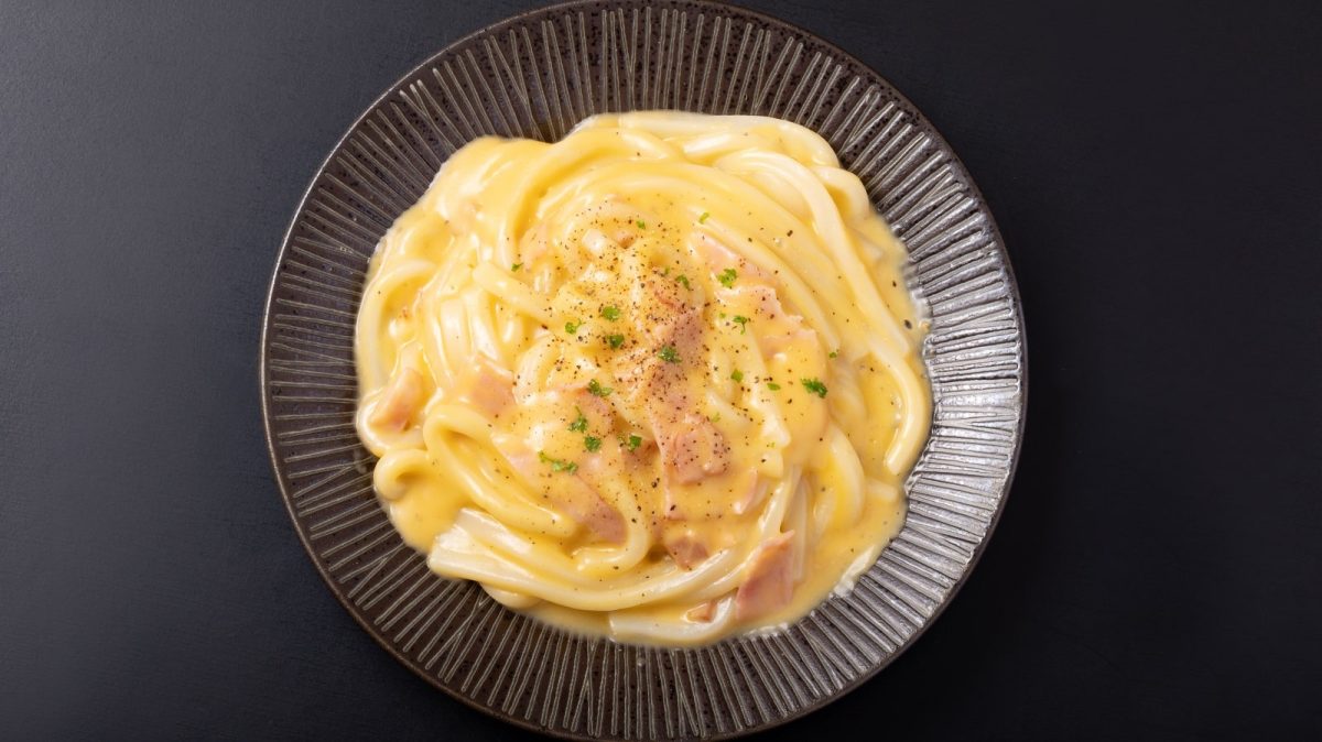 Ein schwarzer Pastateller mit Udon Carbonara auf einem schwarzen Tisch.