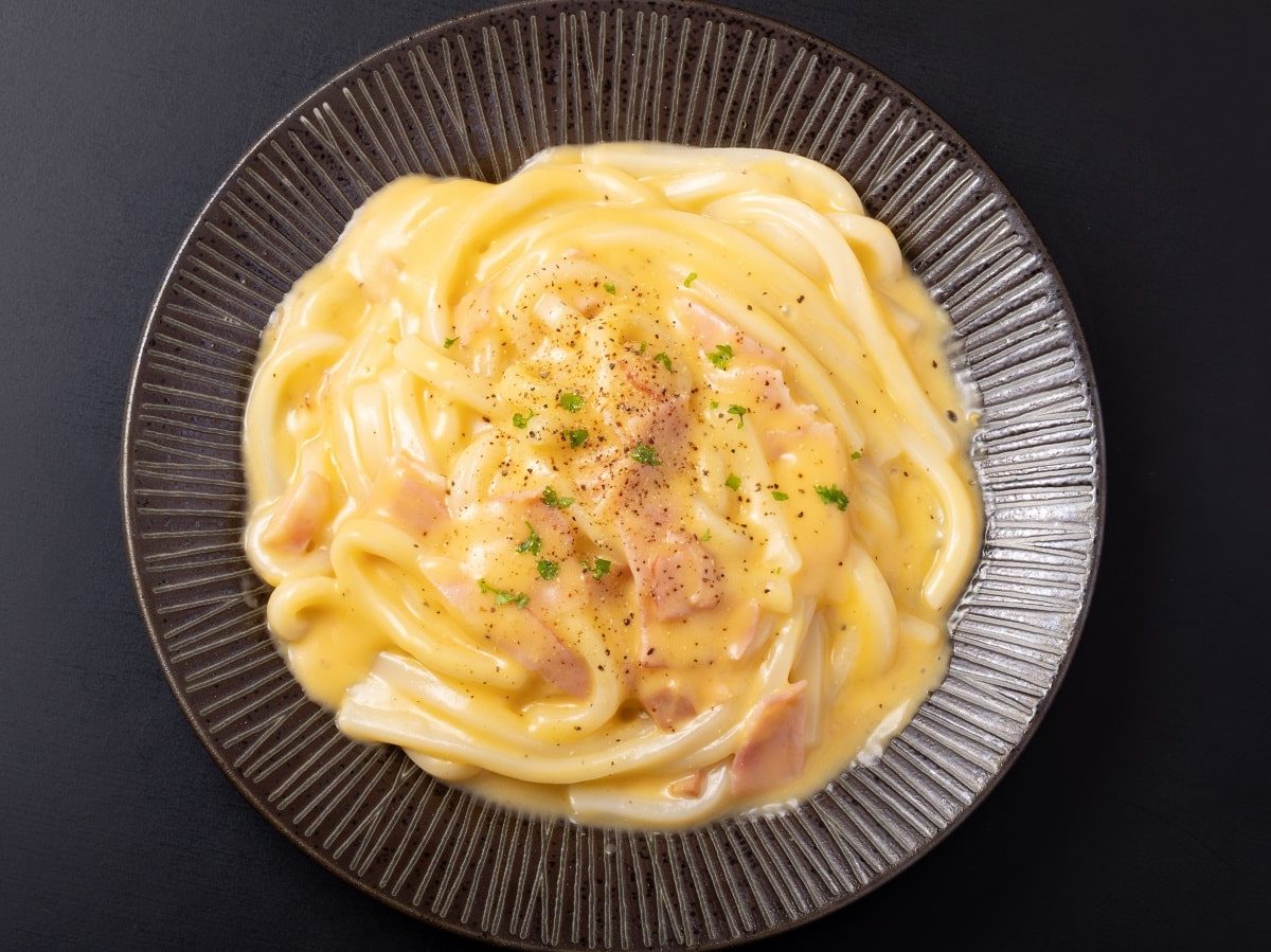 Ein schwarzer Pastateller mit Udon Carbonara auf einem schwarzen Tisch.