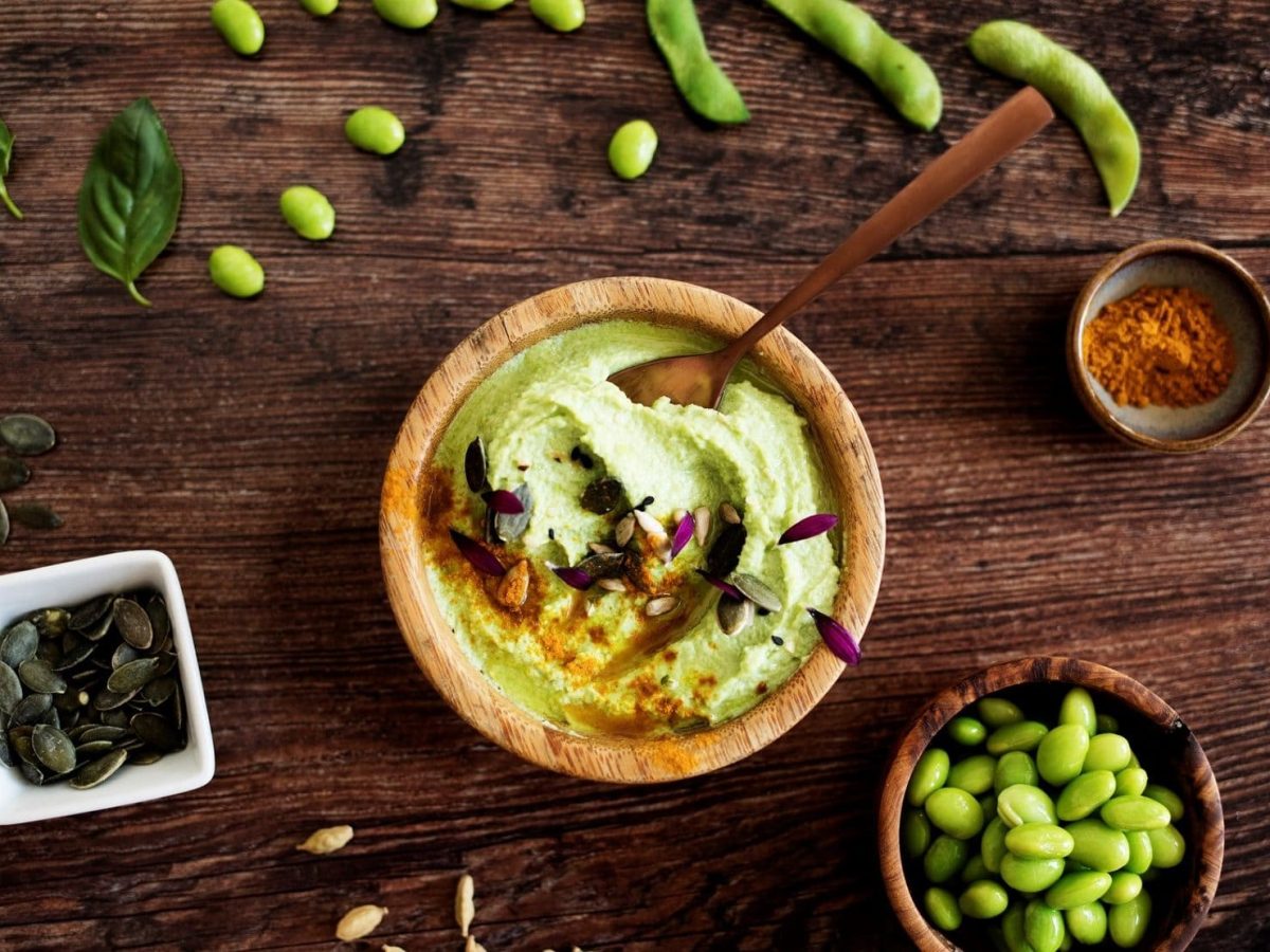 Draufsicht: Schüssel Edamame-Hummus mit Kürbis- und Edamame-Bohnen und Currypulver auf einem Holztisch.