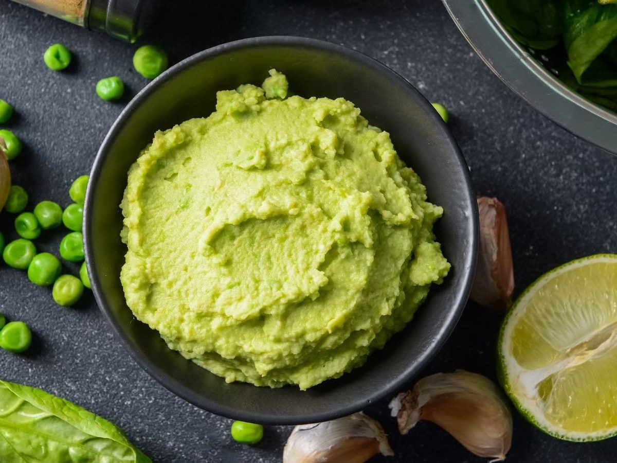 Eine schwarze Schale mit Erbsen-Hummus neben frischen Erbsen.