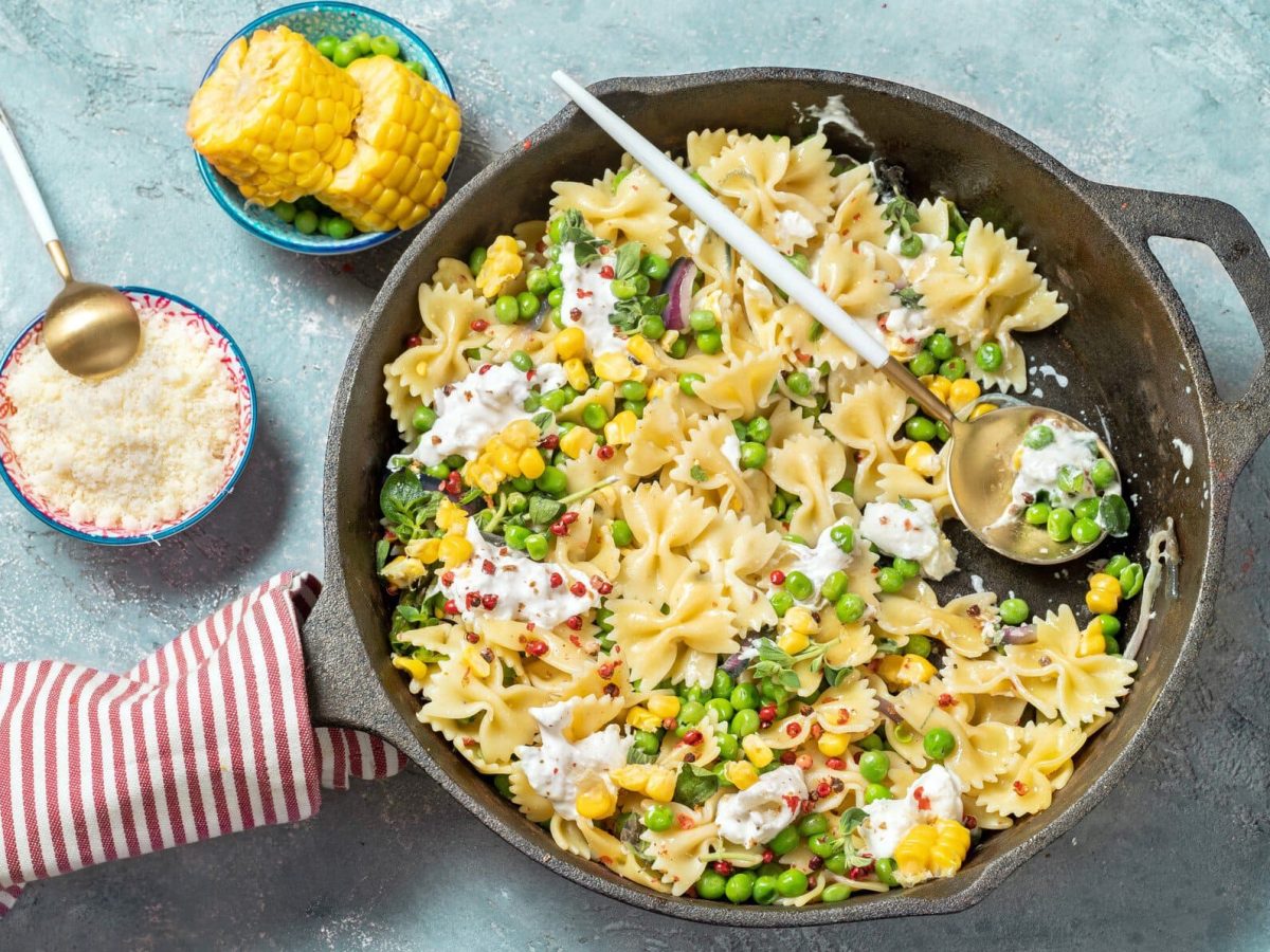 Draufsicht: vegetarische Farfalle mit Erbsen und Burrata auf einen hellblauen Untergrund. Daneben liegen eine Schüssel mit gerieben Parmesan sowie Mais