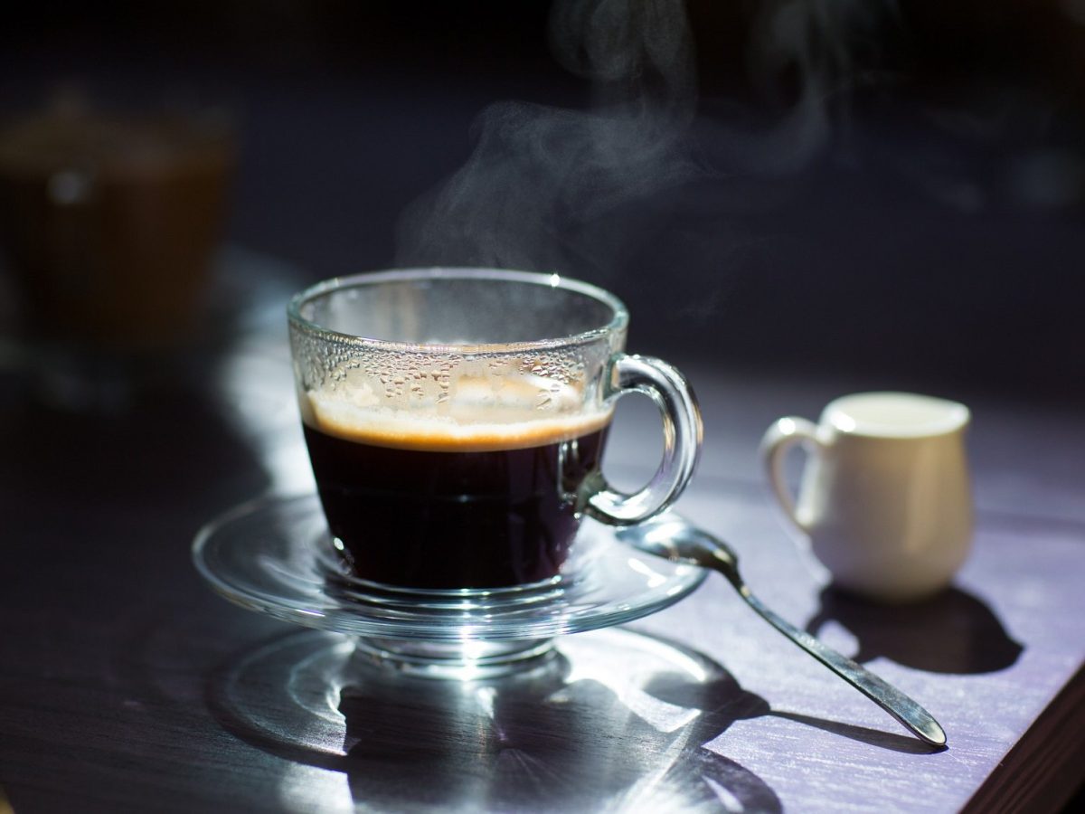 Frontalsicht: Auf auf einem dunklen Tisch stehen vor einem dunklen Hintergrund ein Glas schwarzer Kaffee sowie ein helles weißen Kännchen aus Porzellan.