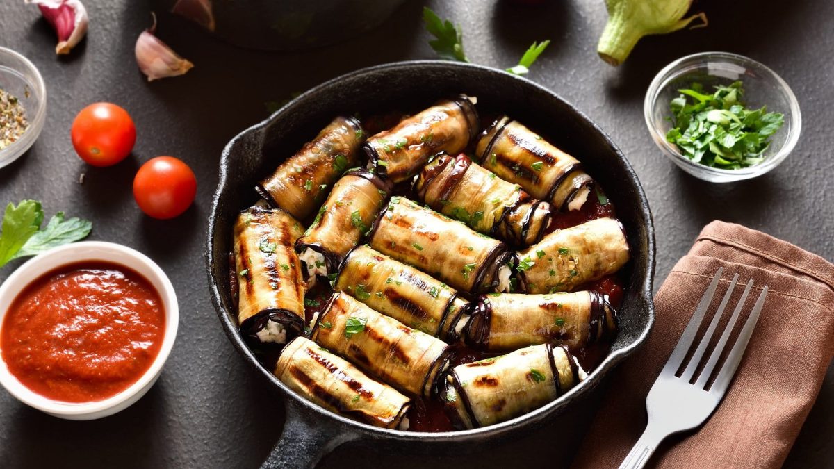 Draufsicht, schräg: Auberginen-Ricotta-Röllchen liegen in einer dunklen Pfanne. Drumherum liegen eine Gabel, eine Schüssel Tomatensauce, Aubergine und Tomaten.