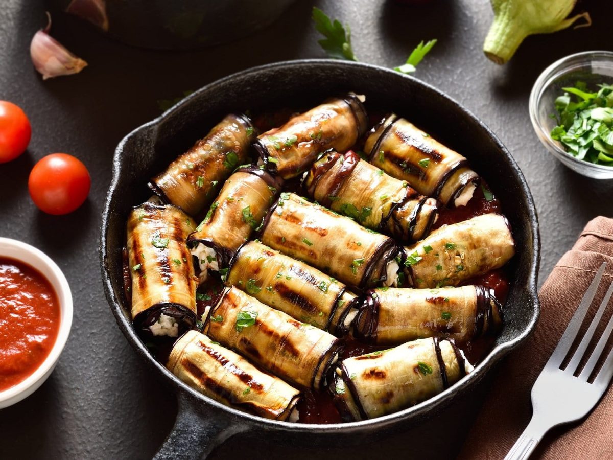 Draufsicht, schräg: Auberginen-Ricotta-Röllchen liegen in einer dunklen Pfanne. Drumherum liegen eine Gabel, eine Schüssel Tomatensauce, Aubergine und Tomaten.