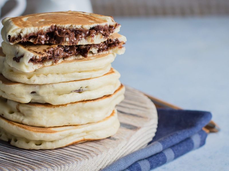 Gefüllte Pancakes mit Nutella gestapelt auf Holzbrett auf Küchentuch. Im Hintergrund eine Tasse. Frontalansicht.