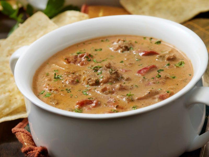 Weiße Schüssel gefüllt mit tomatiger Hackfleisch-Käse-Suppe. Daneben liegen Chips.