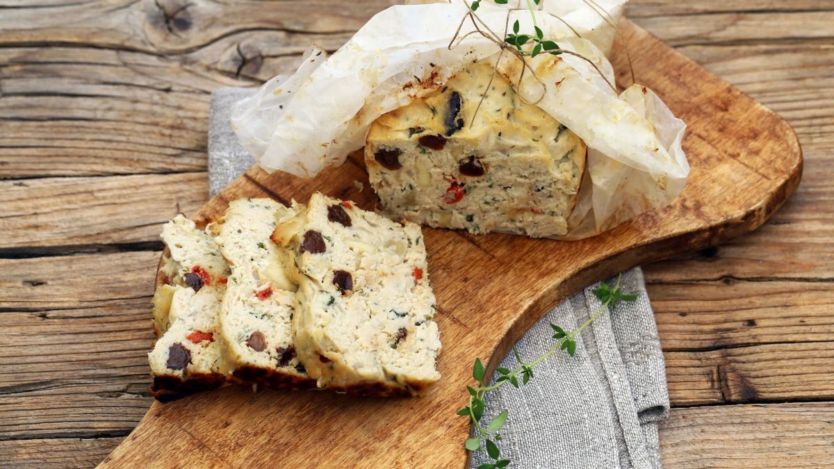 Draufsicht: eine angeschnittene Hähnchen-Terrine mit Tomaten und Oliven auf einem Holzbrettchen.