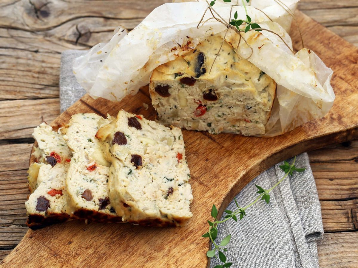 Draufsicht: eine angeschnittene Hähnchen-Terrine mit Tomaten und Oliven auf einem Holzbrettchen.