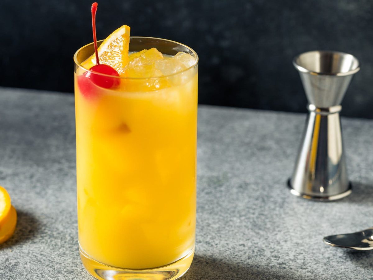 Harvey Wallbanger Cocktail in Longdrinkglas auf grauem Untergrund. Im Bild Barmaß, Barlöffel und Orangenscheibe. Frontalansicht.