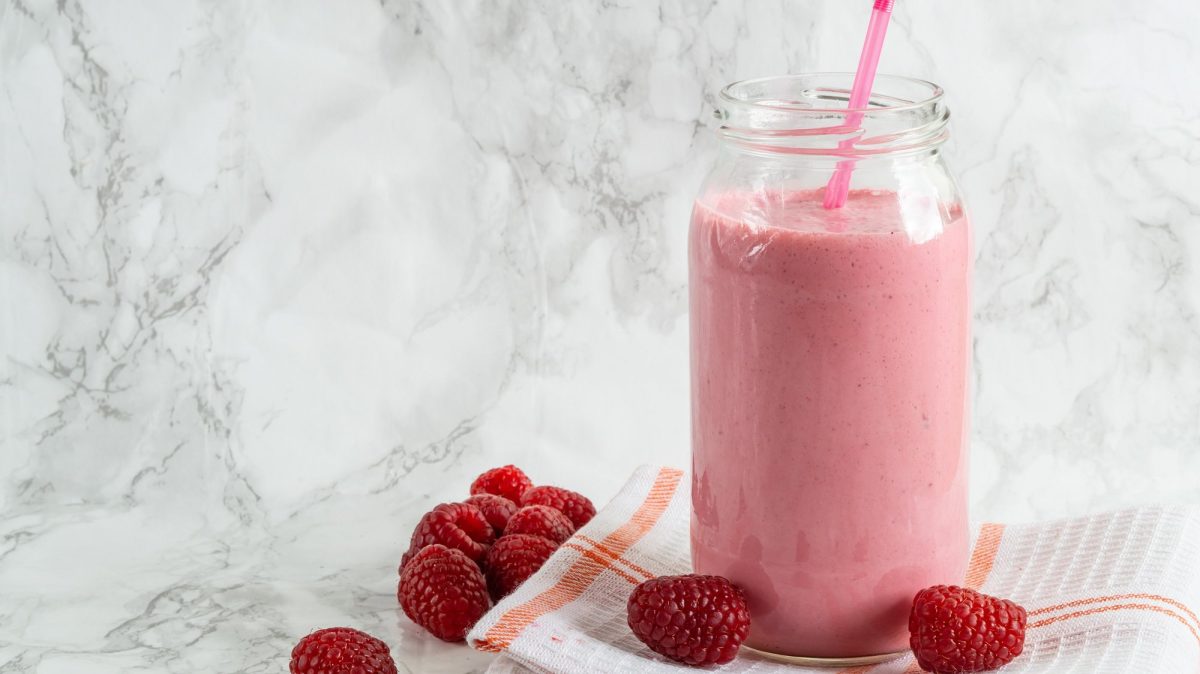 Ein Glas mit Himbeer-Joghurt-Drink und einem Strohhalm, das auf einem Geschirrtuch steht, daneben liegen frische Himbeeren.