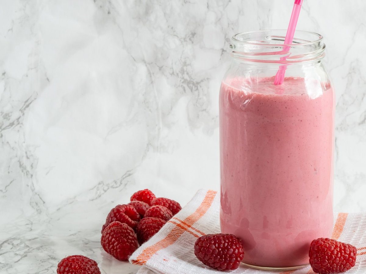 Ein Glas mit Himbeer-Joghurt-Drink und einem Strohhalm, das auf einem Geschirrtuch steht, daneben liegen frische Himbeeren.