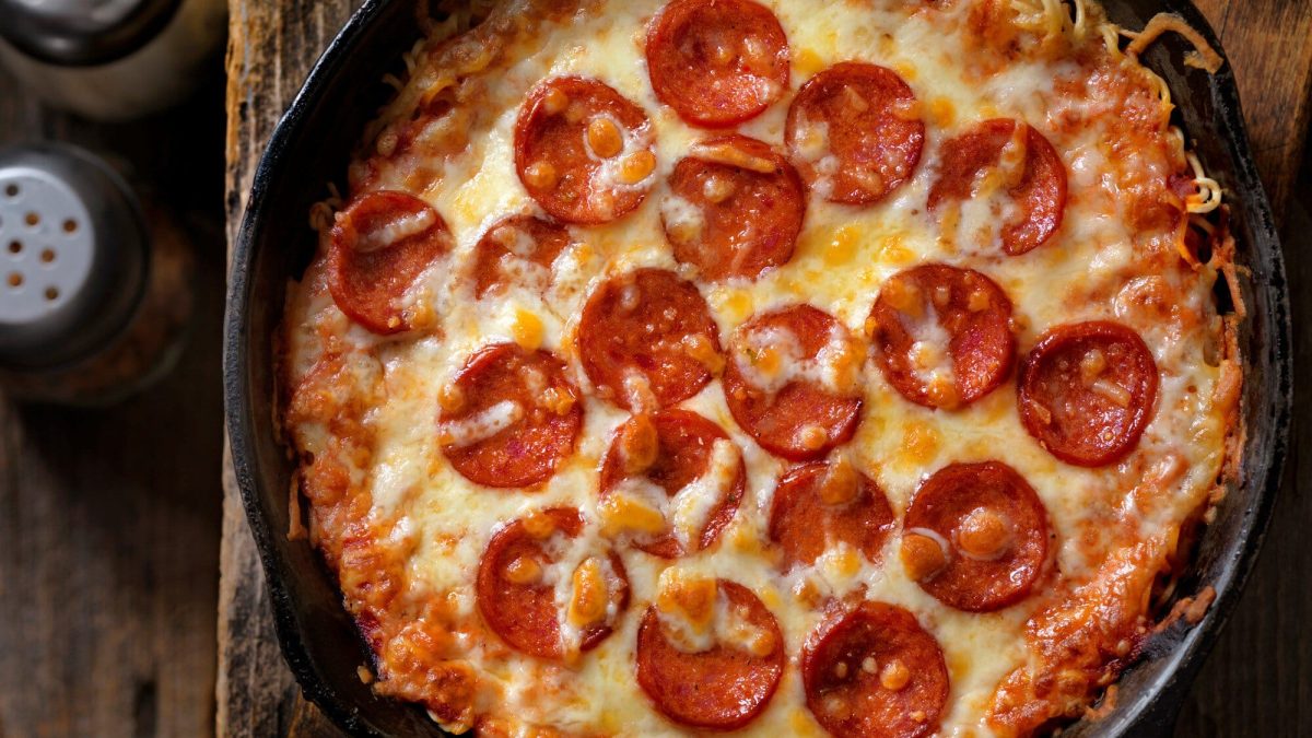 Instant Ramen Pizza in schwarzer Gusspfanne auf Holz. Daneben Salz- und Pfefferstreuer. Draufsicht.