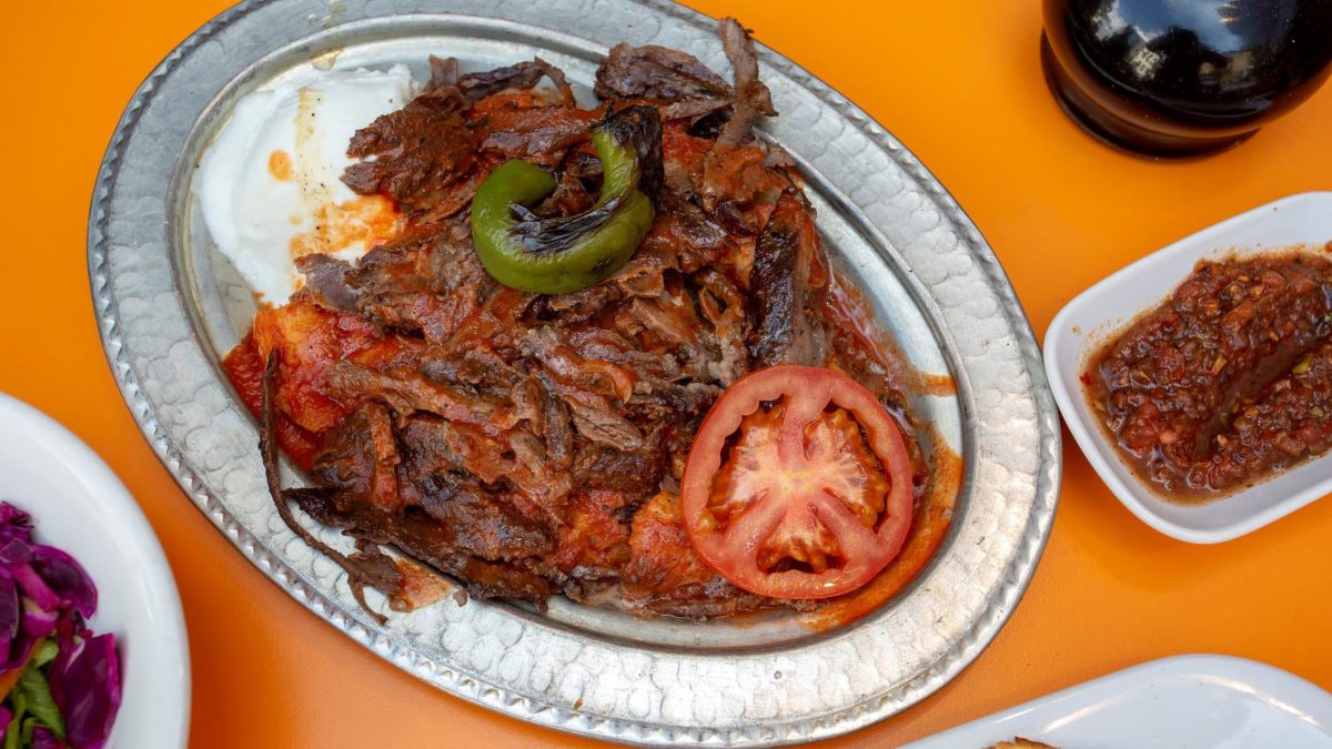 Iskender Kebab angerichtet auf Platte auf orangenen Tisch. Im Bild weitere Zutaten. Draufsicht.