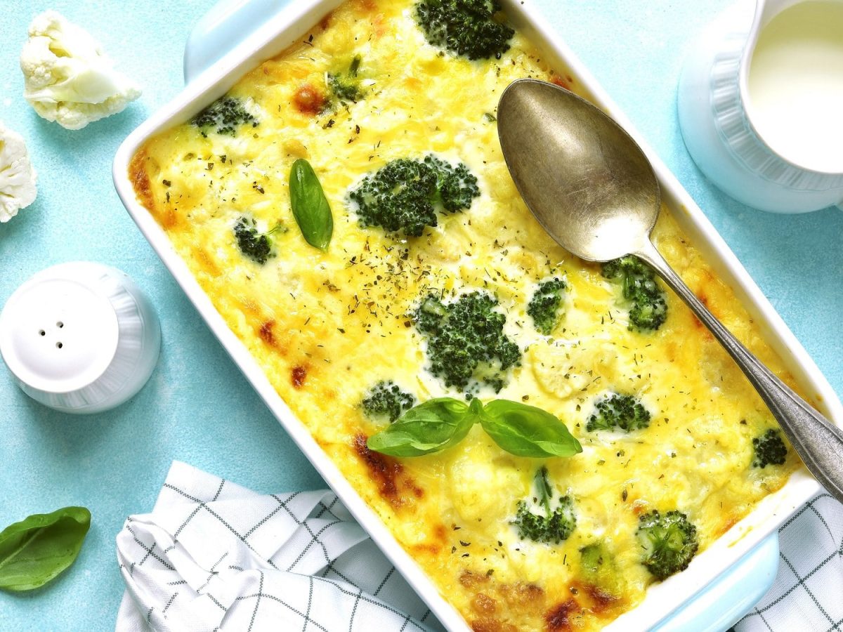 Draufsicht: Auf einem hellblauem Untergrund steht ein Brokkoli-Gratin mit Blumenkohl. Drumherum liegen Basilikumblätter und ein Kännchen mit Sahne sowie ein Salzstreuer