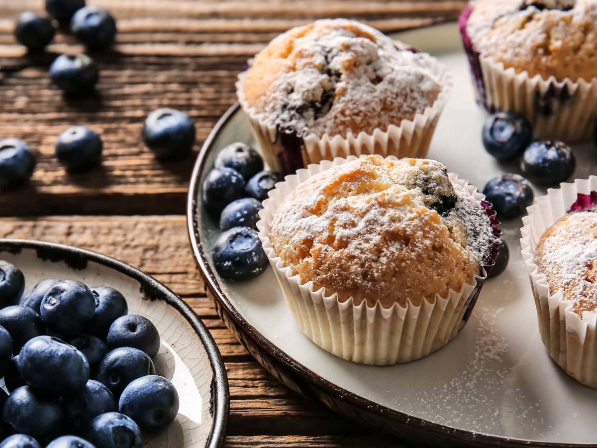 Draufsicht: Muffins mit Beeren stehen auf einem Teller, der auf einem Holztisch liegt. Daneben ist ein kleiner Teller mit Blaubeeren. Auch auf den Tisch liegen noch frische Beeren.