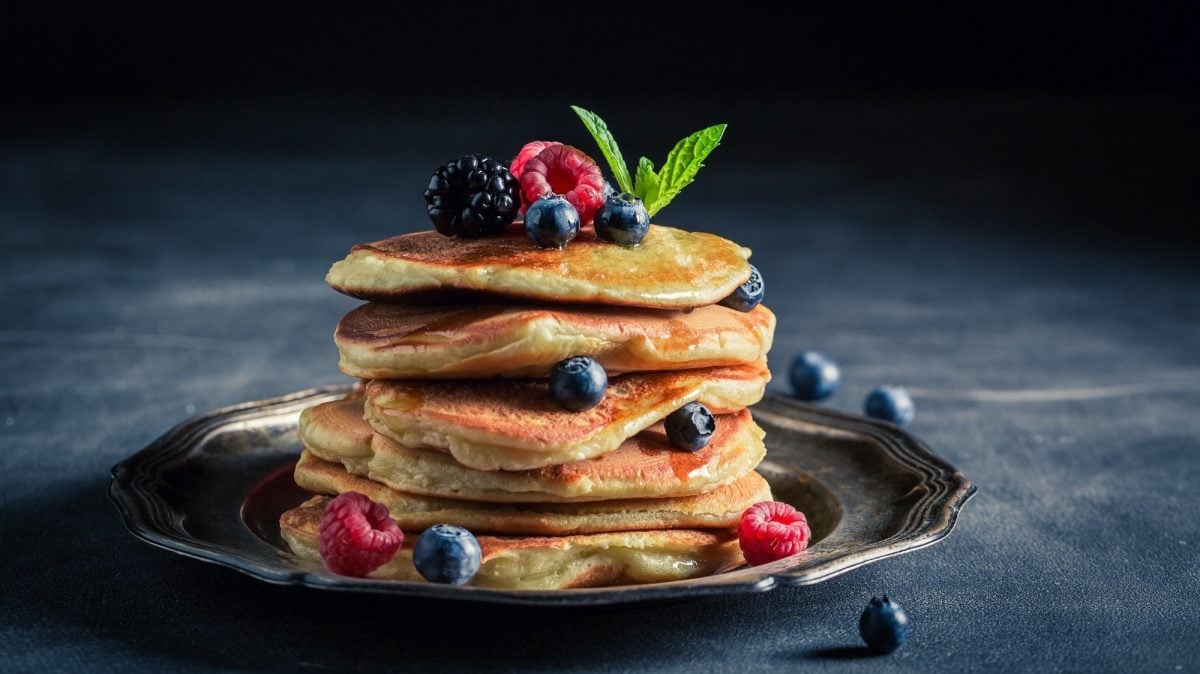 Ein Stapel Joghurt-Zitronen-Pancakes auf einem Teller, garniert mit verschiedenen frischen Beerensorten und Minze.