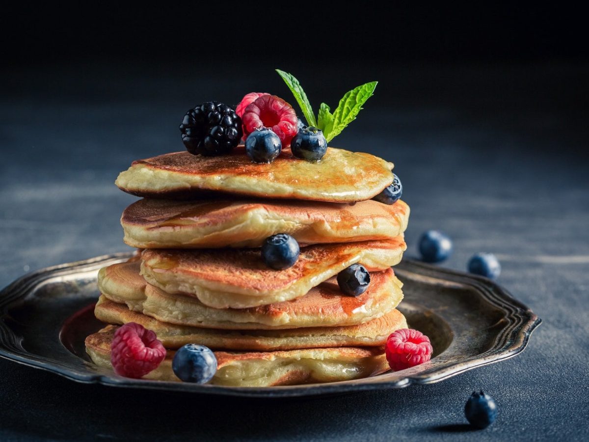 Ein Stapel Joghurt-Zitronen-Pancakes auf einem Teller, garniert mit verschiedenen frischen Beerensorten und Minze.