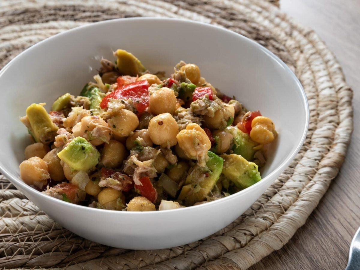 Kichererbsensalat mit Thunfisch und Avocado in weißer Schale auf Untersetzer und Holztisch. Daneben eine Gabel. Draufsicht.