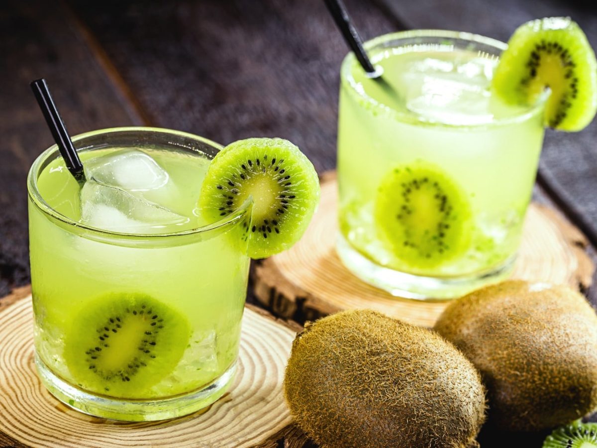 Zwei grüne Cocktails mit Kiwi auf zwei Holzscheiben und dunklem Untergrund.