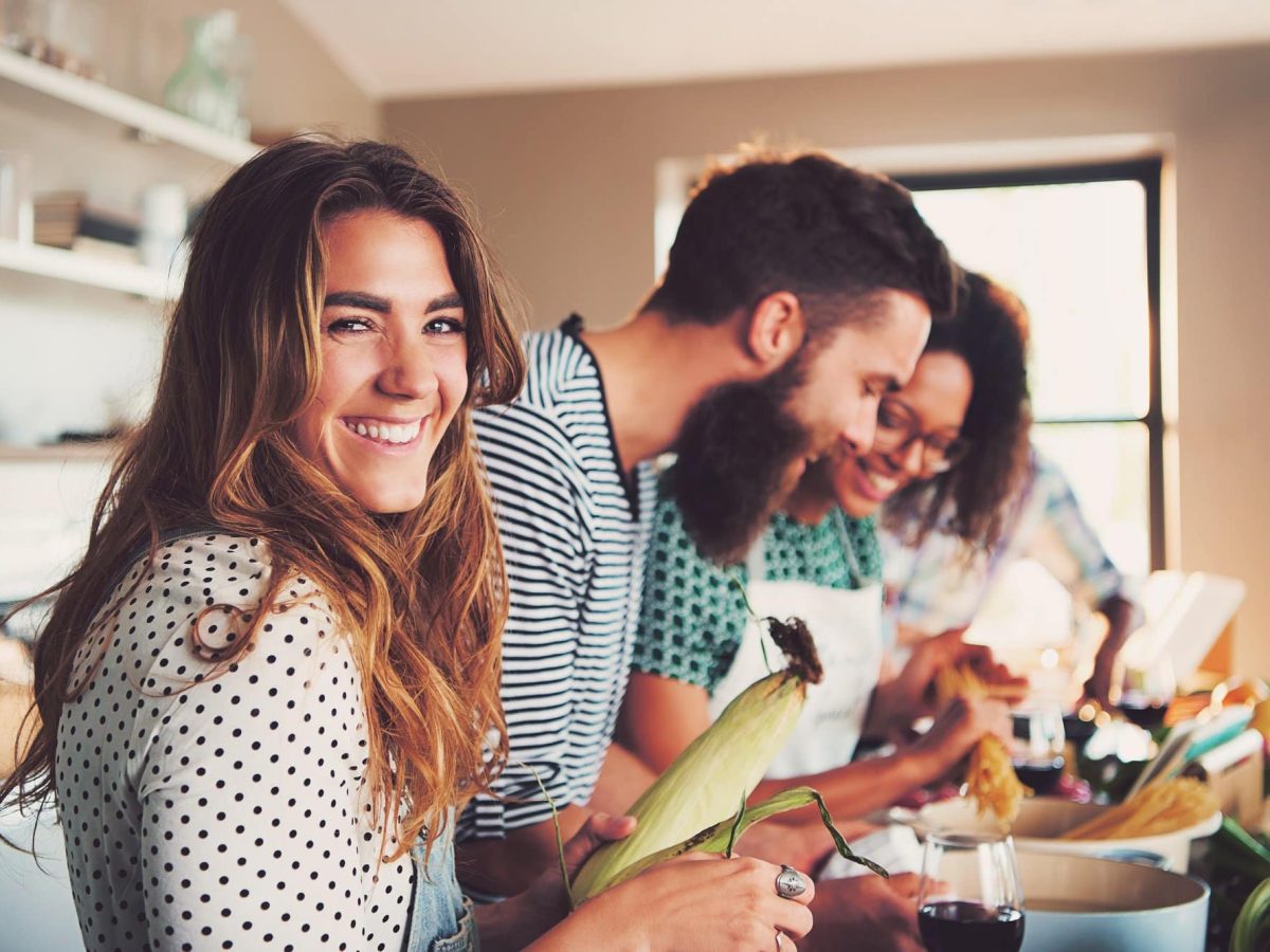 Kochen ohne Rezepte: Eine Gruppe von Freunden in der Küche beim Kochen, eine Frau lacht in die Kamera.