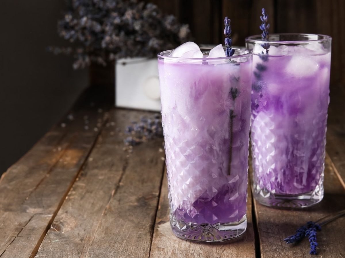 Zwei Gläser mit Lavendel-Cocktail mit Likör 43 mit Eiswürfeln und Strohhalmen auf einem Holztisch vor dunklem Hintergrund.