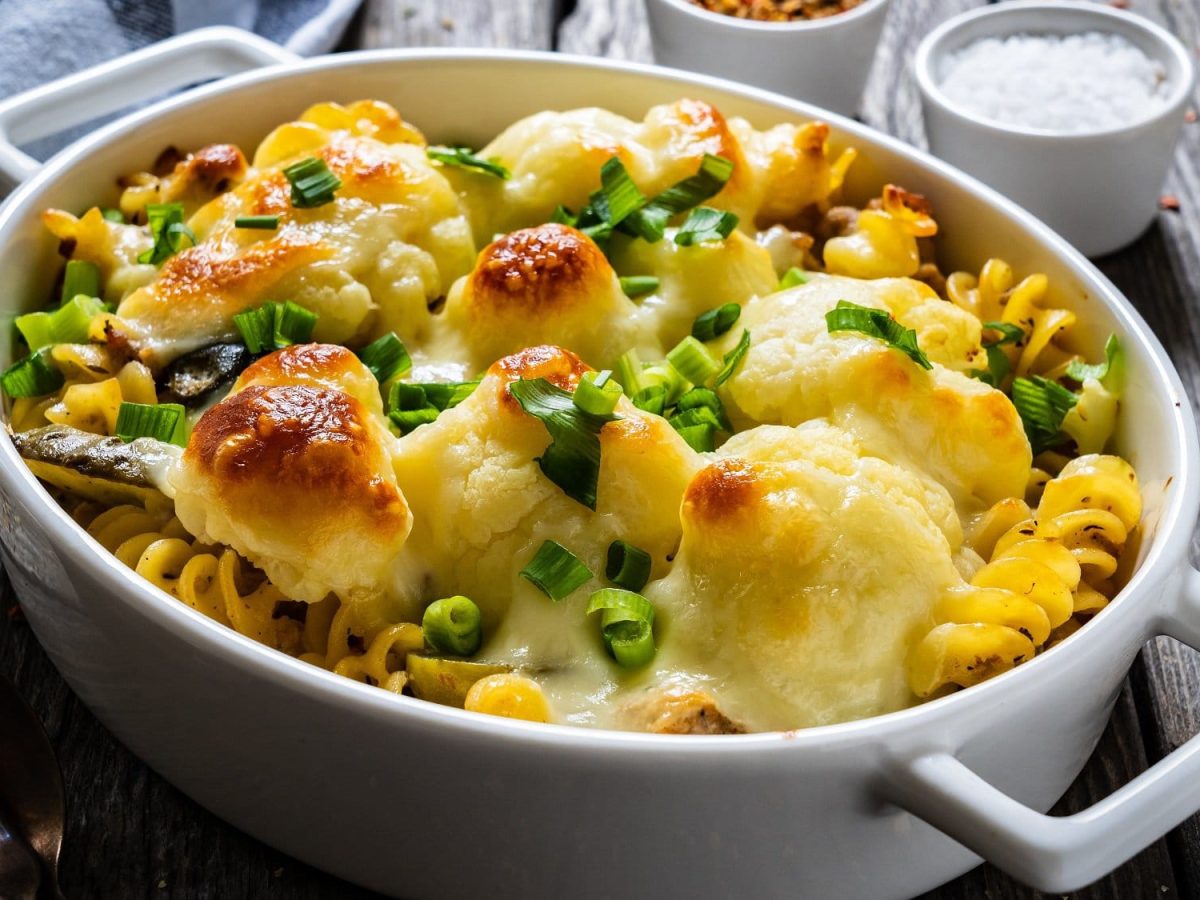 Schräge Draufsicht: Eine weiße Form mit einem Blumenkohl-Hackauflauf der mit Käse überbacken wurde. Der steht auf einem dunklen Holztisch. Daneben liegen mehrere kleine Gefäße mit Gewürzen, wie etwa Salz und Pfeffer, sowie zwei Gabeln und ein Küchentuch.