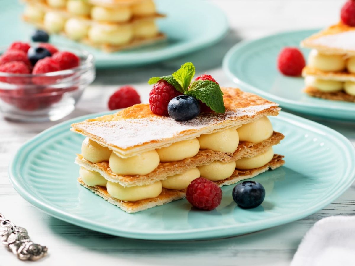 Drei blaue Teller mit dem Mille-feuille auf einem hellen Holztisch mit eine rweißen Serviette und einer silbernen Gabel.