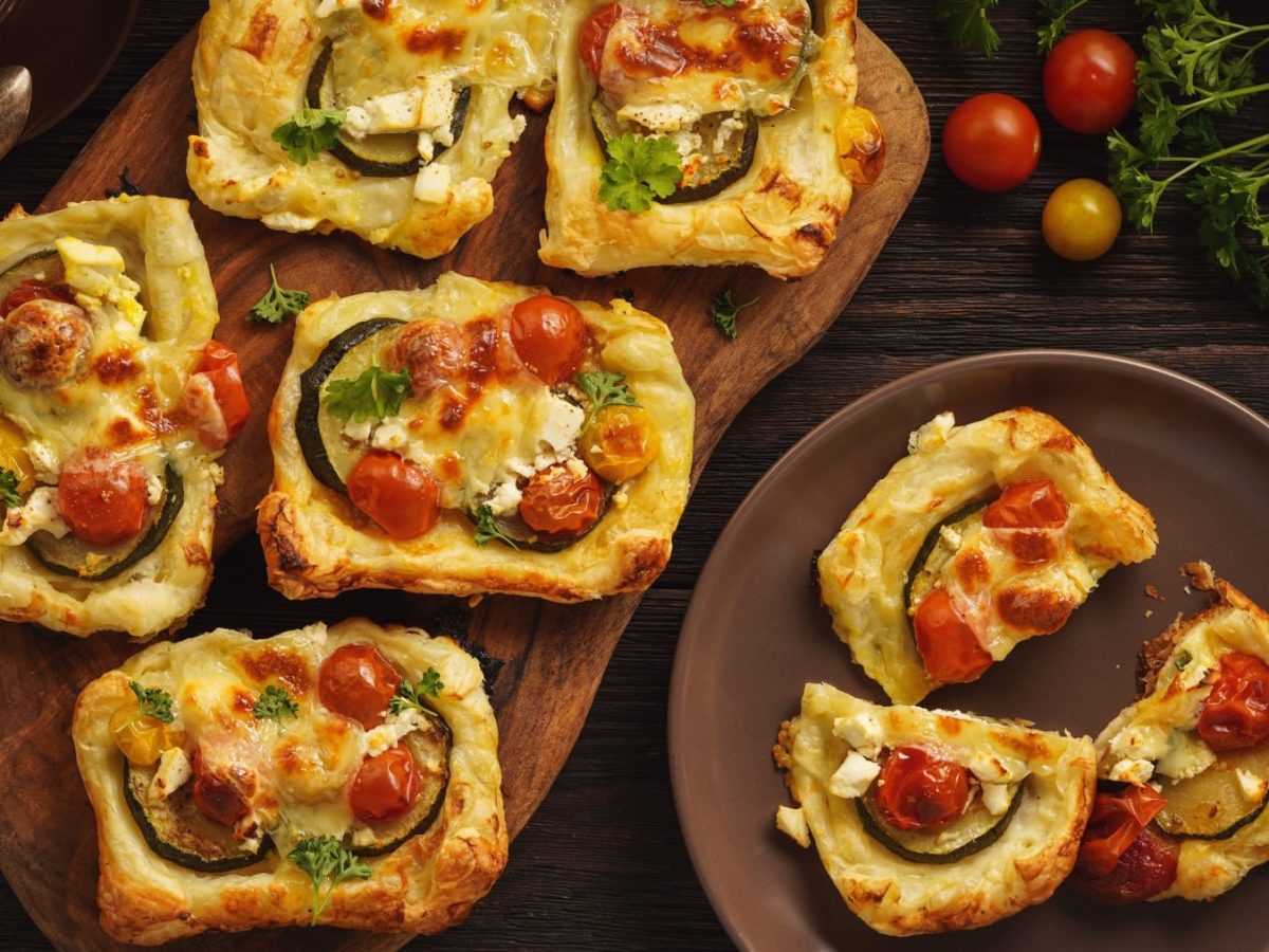 Mehrere Mini-Blätterteig-Tartes mit Zucchini, Tomaten und Feta in der Draufsicht.