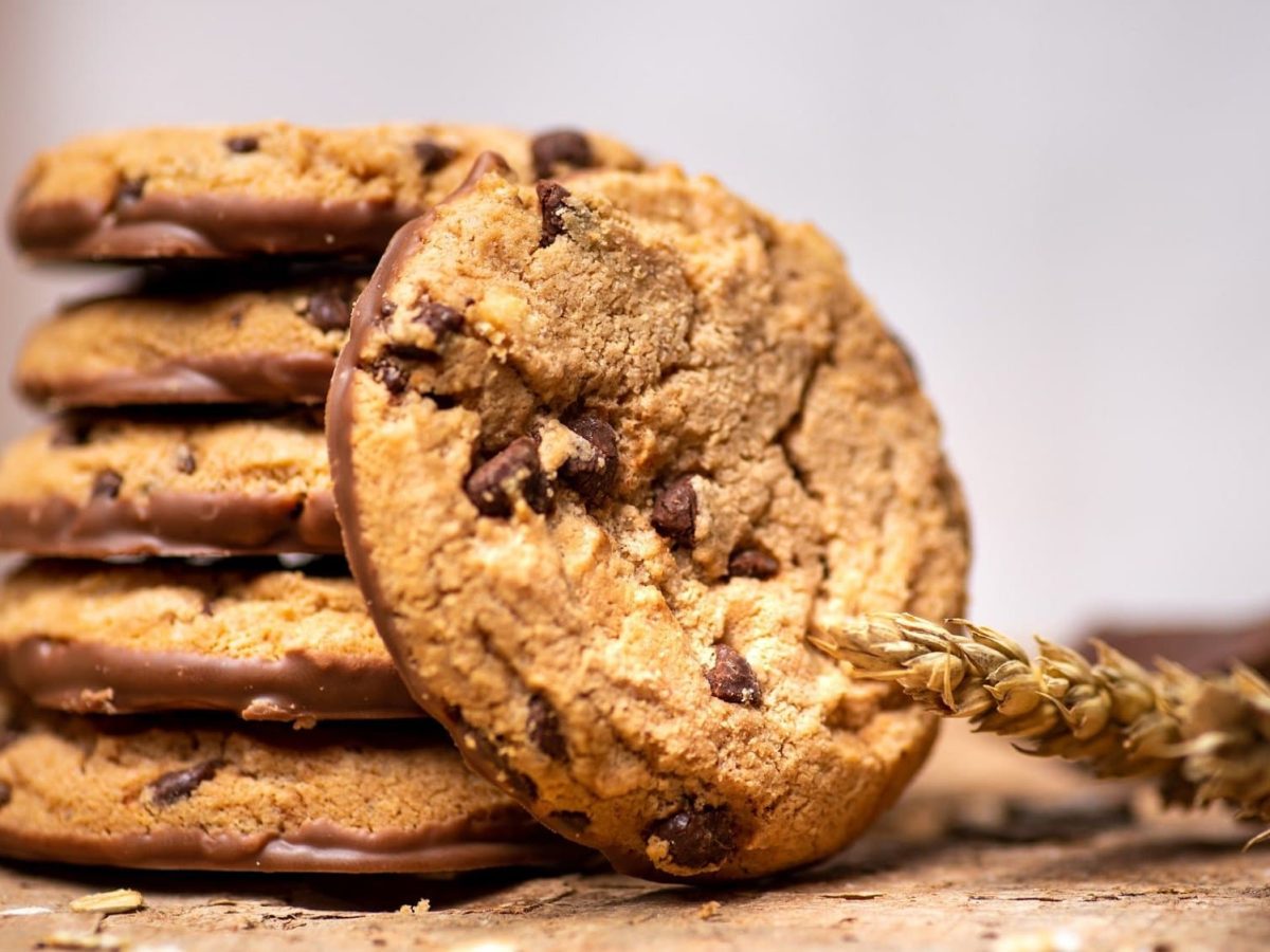 Frontalsicht: An einem Stapel Peanut and Chocolate Cookies, ist ein weiterer Keks angelehnt. Daneben liegt eine Weizenähre.