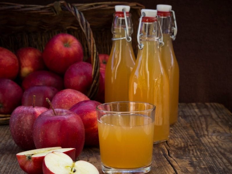 Frontalsicht: Drei Flaschen sekbst gemachten Apfelsaft, sowie mehrere Äpfel und Apfelhälften stehen auf einem Holztisch vor einem dunklen Hintergrund.