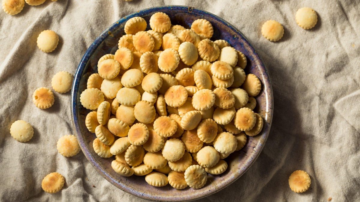 Oyster Cracker in einer grauen Schüssel auf beigem Tuch. Weitere Cracker im Bild verteilt. Draufsicht.