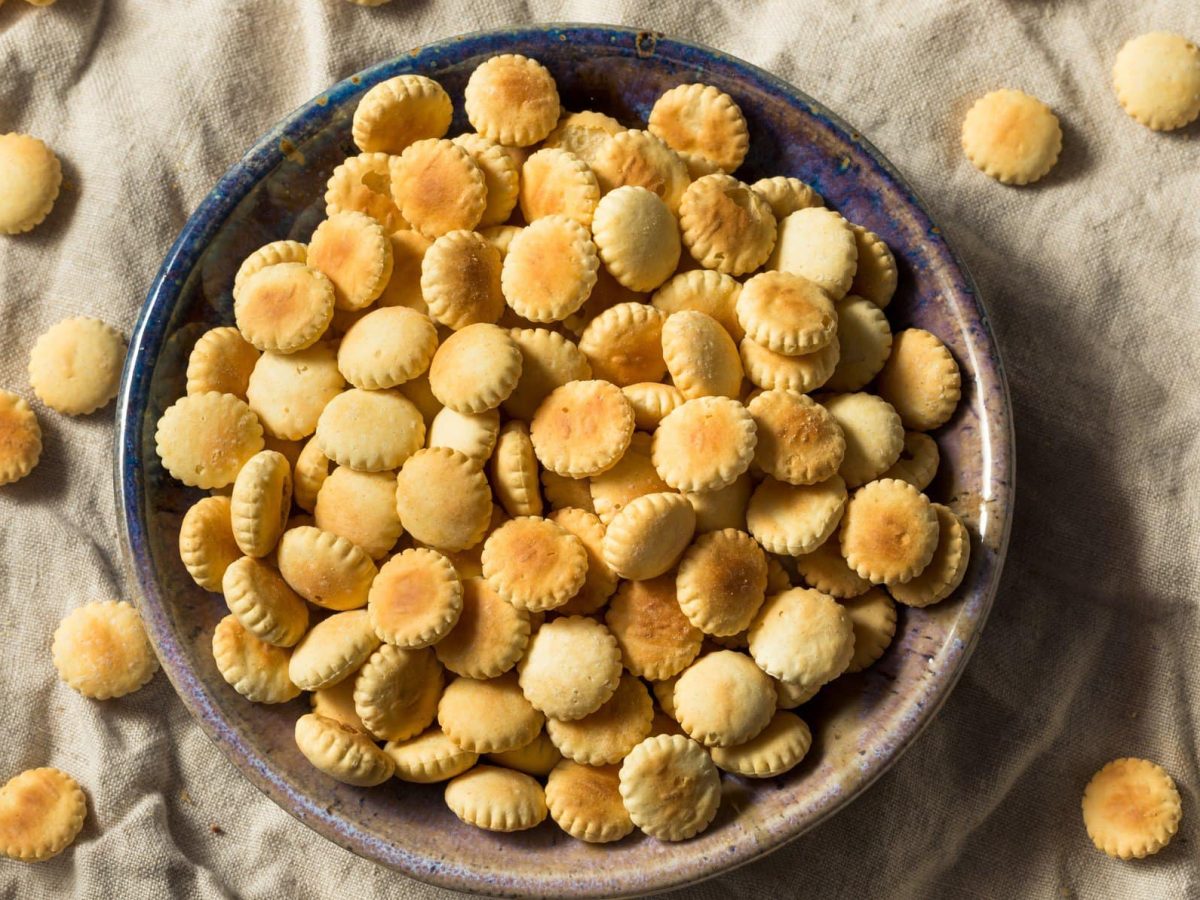 Oyster Cracker in einer grauen Schüssel auf beigem Tuch. Weitere Cracker im Bild verteilt. Draufsicht.