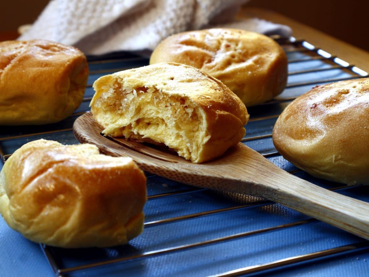 Fünf Pan de Coco auf Rost auf blauem Untergrund. Eine halbiert auf Holzkochlöffel. Draufsicht.