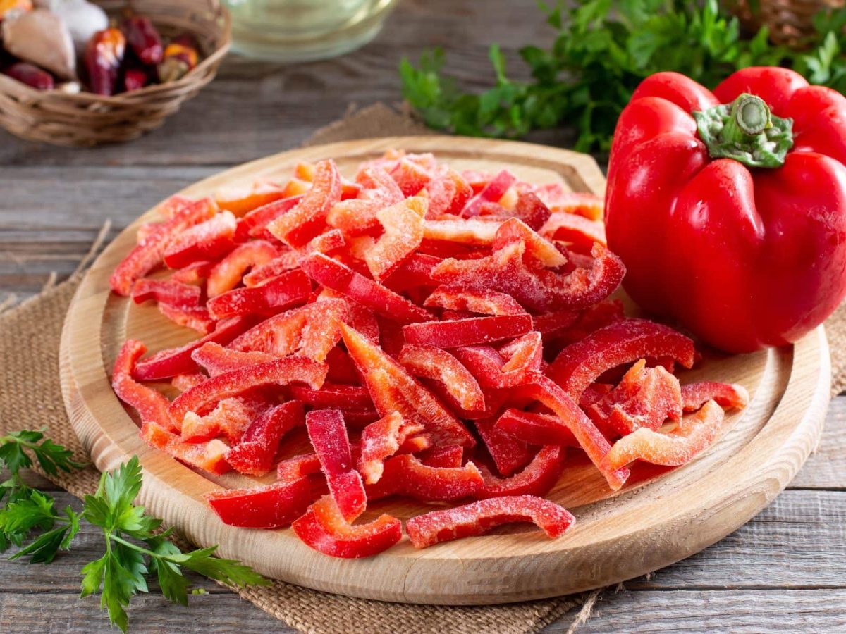 Paprika einfrieren gefrorene Streife Paprika und eine ganze frische rote Paprika auf Holzbrett. Im Hintergrund Kräuter und Schalotten