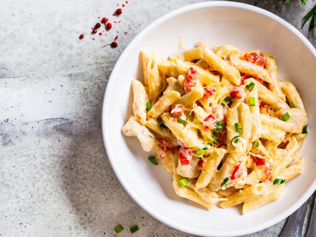 Ein Teller Pasta mit Hummus-Sauce, Frühlingszwiebeln und Paprika in der Draufsicht, daneben Besteck.