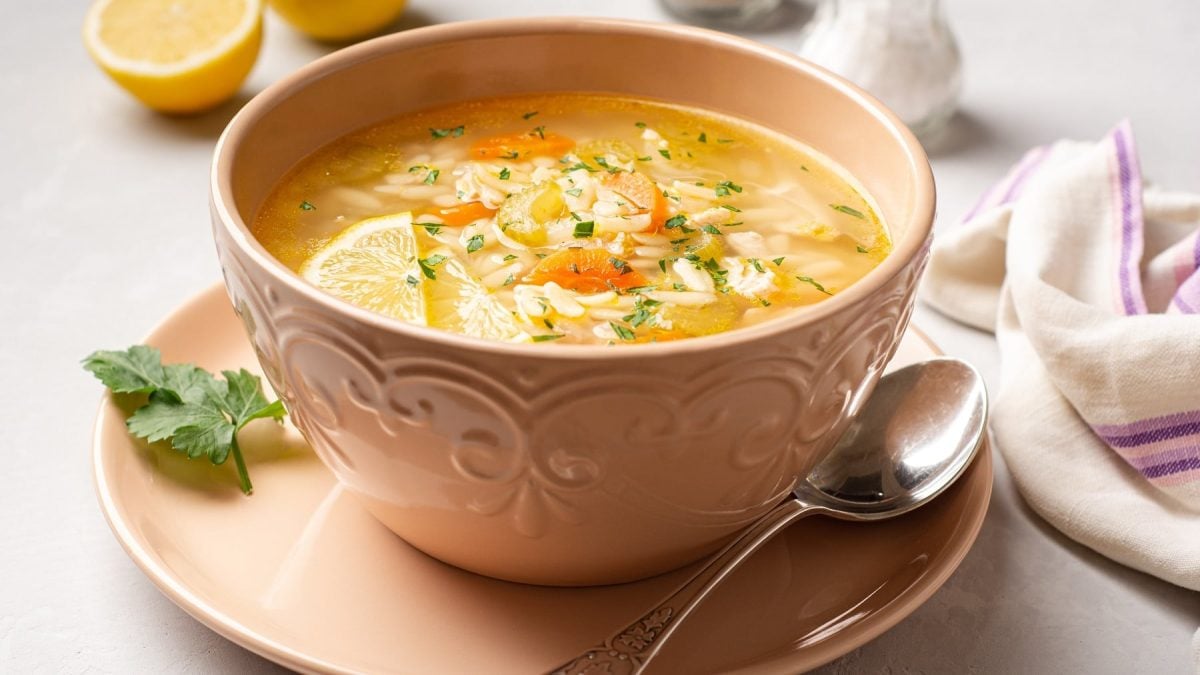 Eine rosa Schale mit der Pastina-Suppe auf einem weißen Tisch mit halbierter Zitrone, Salz- und Pfefferstreuer.