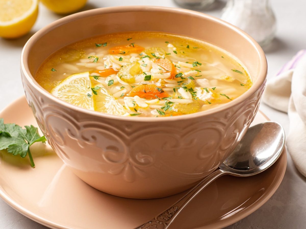 Eine rosa Schale mit der Pastina-Suppe auf einem weißen Tisch mit halbierter Zitrone, Salz- und Pfefferstreuer.