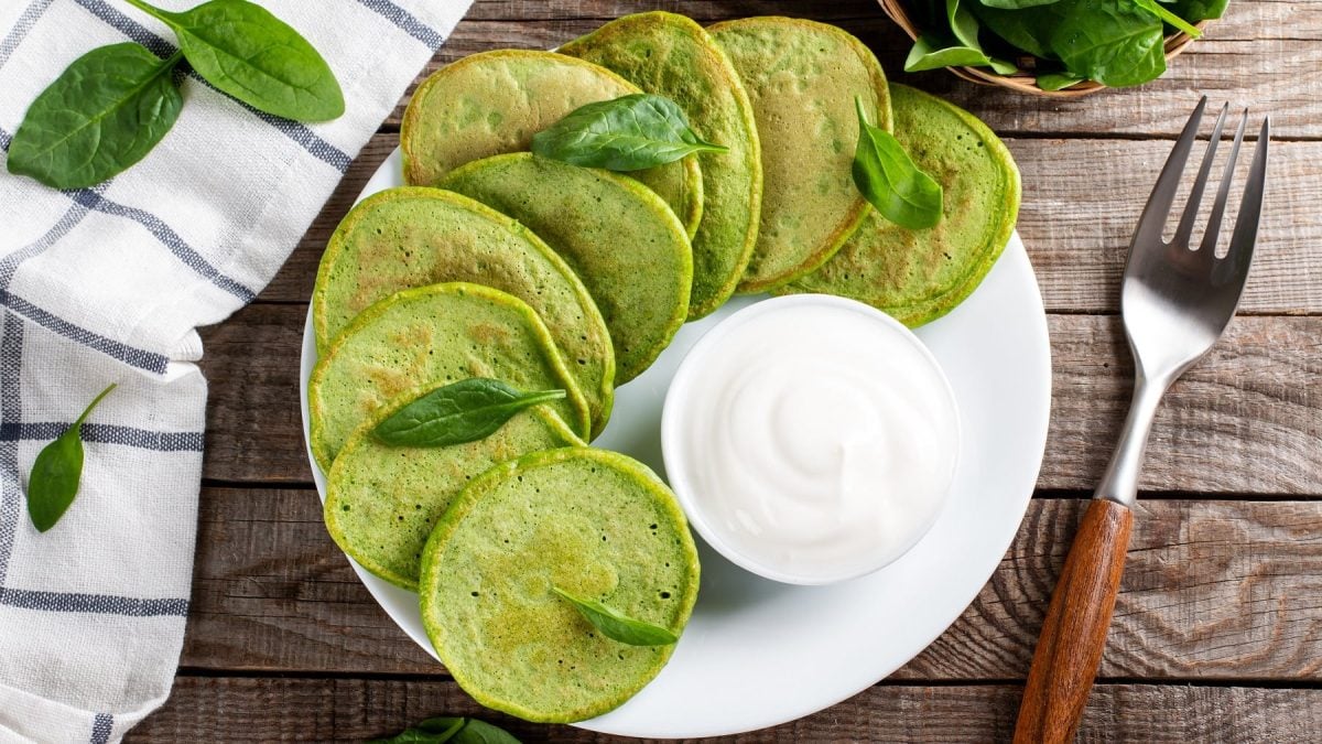 Ein weißer Teller mit den Pesto-Pfannkuchen auf einem Holztisch mit Spinatblättern und einer Gabel.