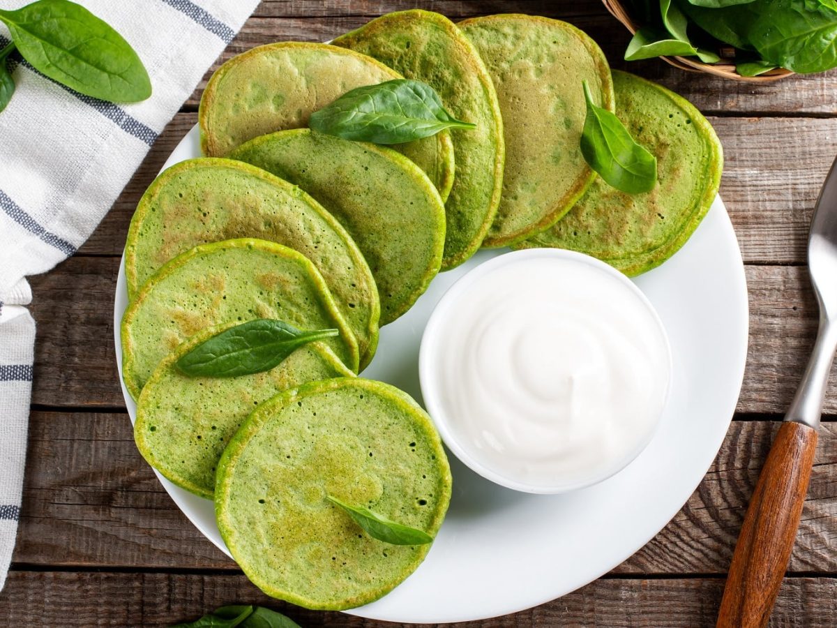 Ein weißer Teller mit den Pesto-Pfannkuchen auf einem Holztisch mit Spinatblättern und einer Gabel.