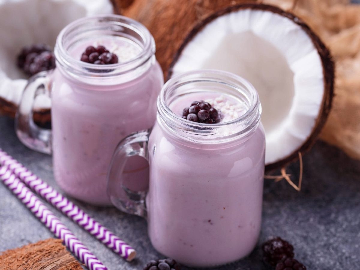 Schräge Draufsicht: Zwei Gläser Kokos-Brombeer-Shake stehen auf einem dunklen Untergund. Daneben liegen zwei geringelte Strohhalme, zwei Kokosnuss-Hälften und Brombeeren.