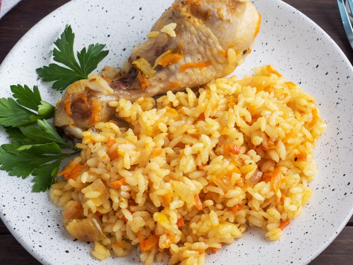 Plov mit Drumstick auf weißem Teller mit Petersilie. Daneben Gabel und Geschirrtuch. Draufsicht.