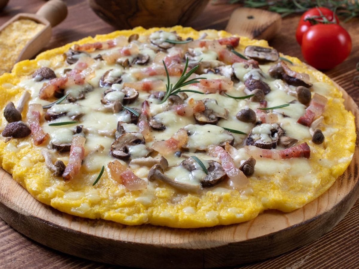 Die Polenta-Pizza auf einem runden Holzschneidebrett. Daneben Maisgrieß, Kirschtomaten und Pilze.