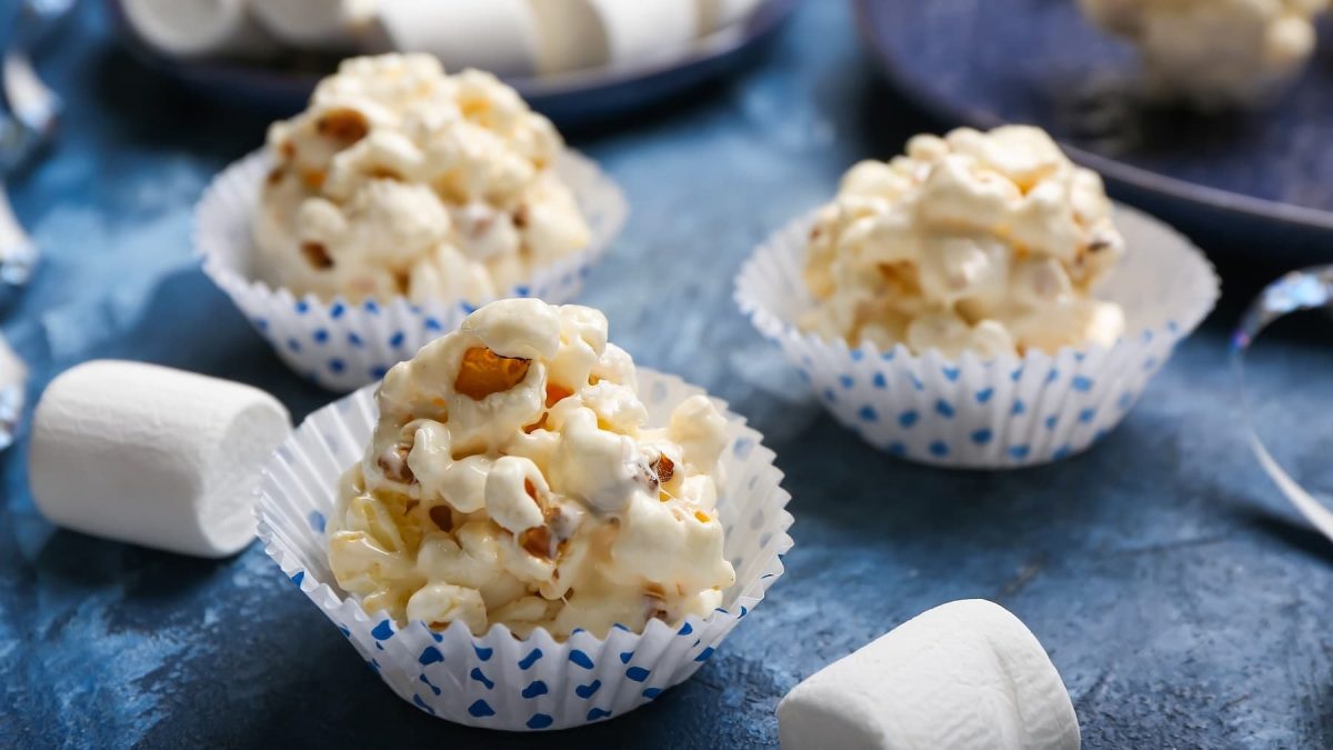 Drei Muffinförmchen mit den Popcorn-Balls und einigen Marshmallows auf einem blauen Tisch.