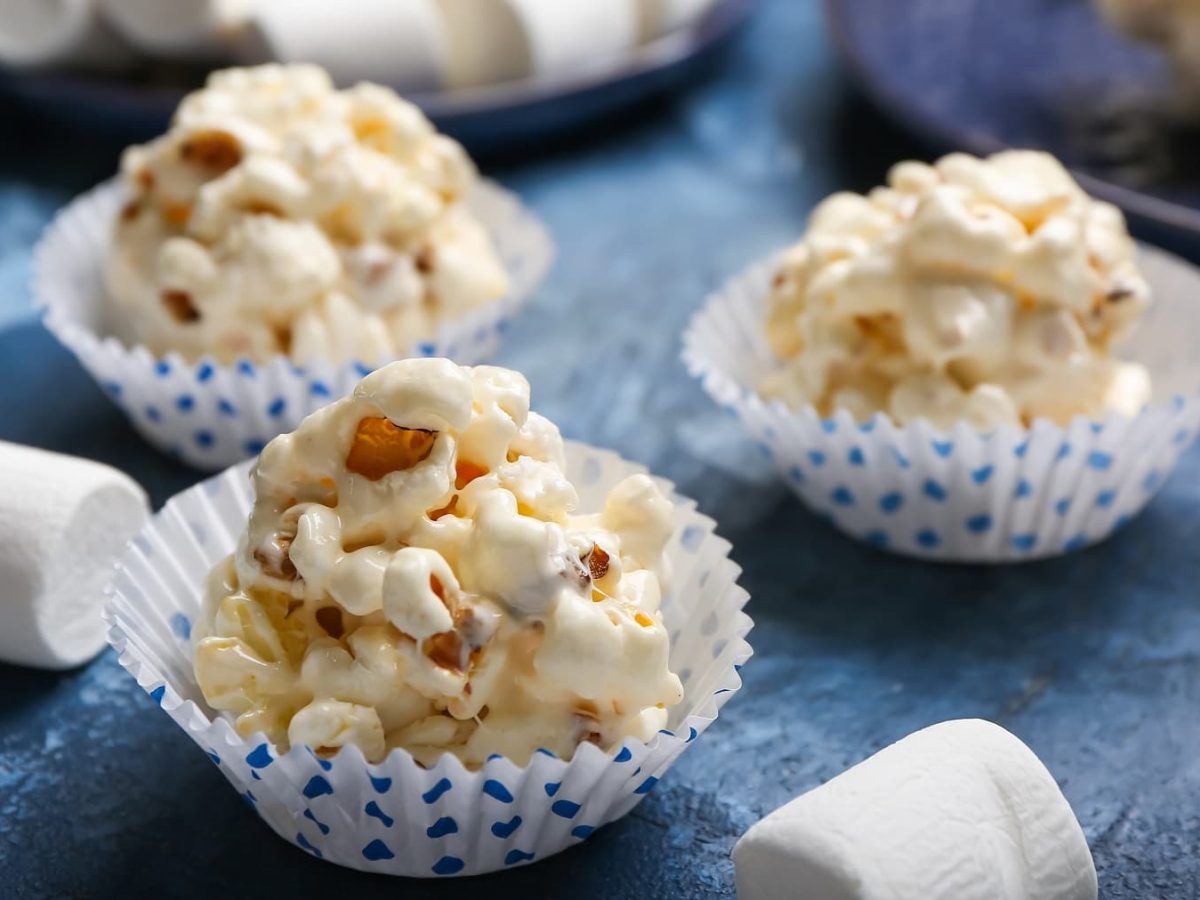 Drei Muffinförmchen mit den Popcorn-Balls und einigen Marshmallows auf einem blauen Tisch.